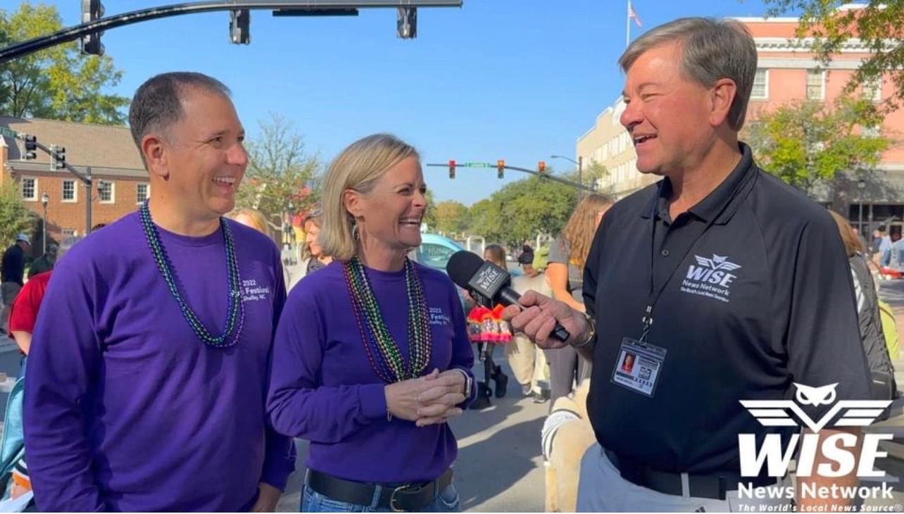 Two people wearing purple shirts and beads being interviewed by a man in a black shirt from WISE News Network at an outdoor event.