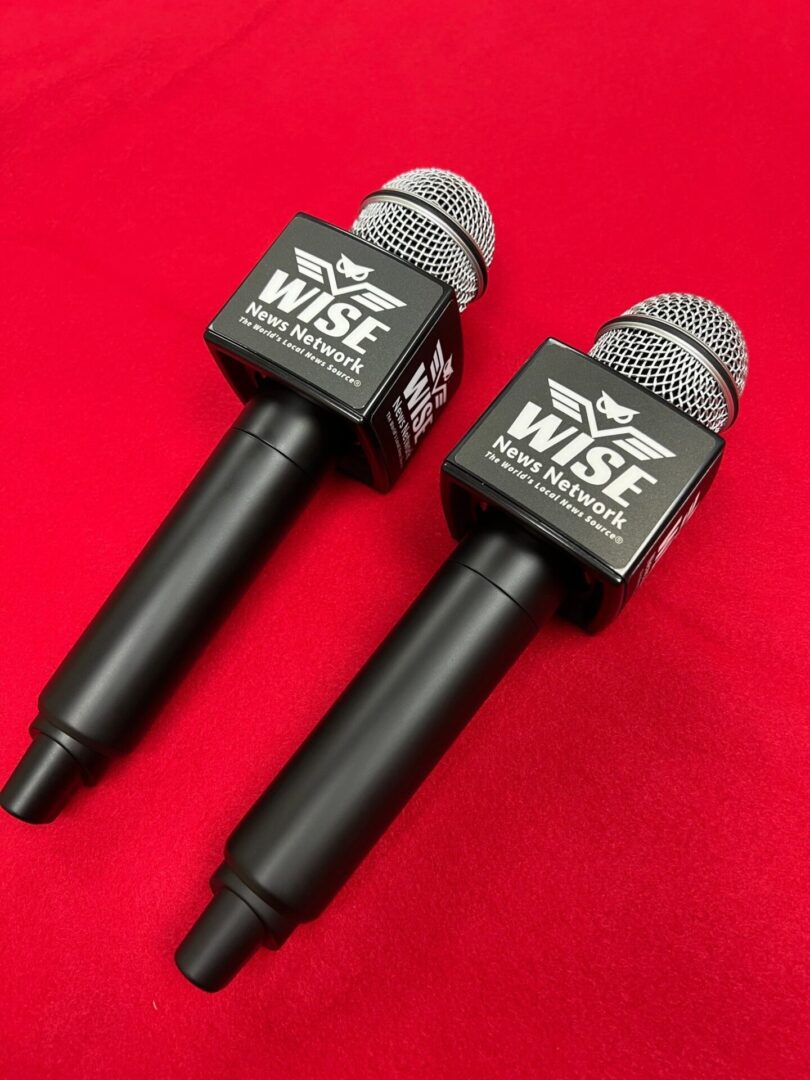 Two black handheld microphones with foam windscreens and logo stickers that read "WISE News Network" placed on a red surface.