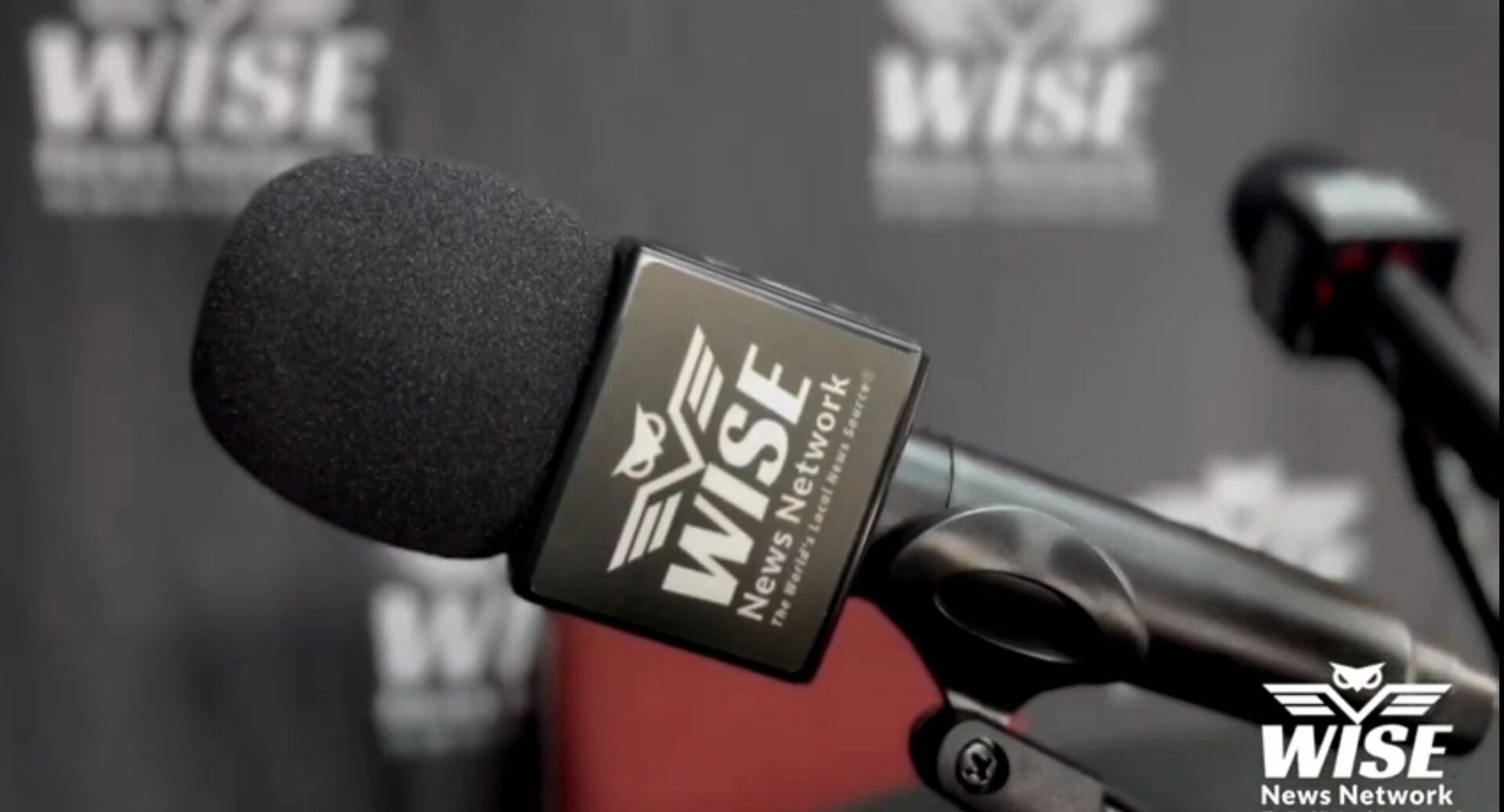 Close-up of a microphone with the WISE News Network logo on it, set against a backdrop displaying the same WISE News Network branding.