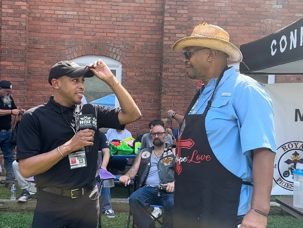 Two men in conversation outdoors; one holds a microphone and wears a black outfit, the other wears a straw hat and apron with "Hope Love" on it. A brick wall and seated individuals are visible in the background.