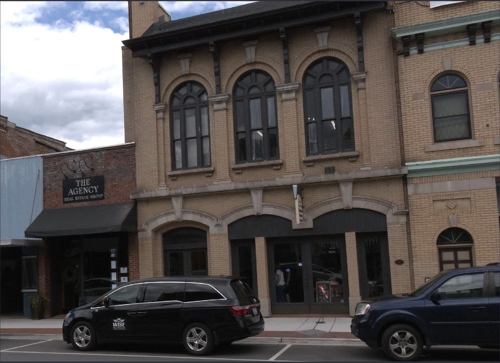 A street with two parked cars and a row of buildings, including one with a wooden sign reading "The Agency Real Estate Group." The buildings have arched windows and brick exteriors.