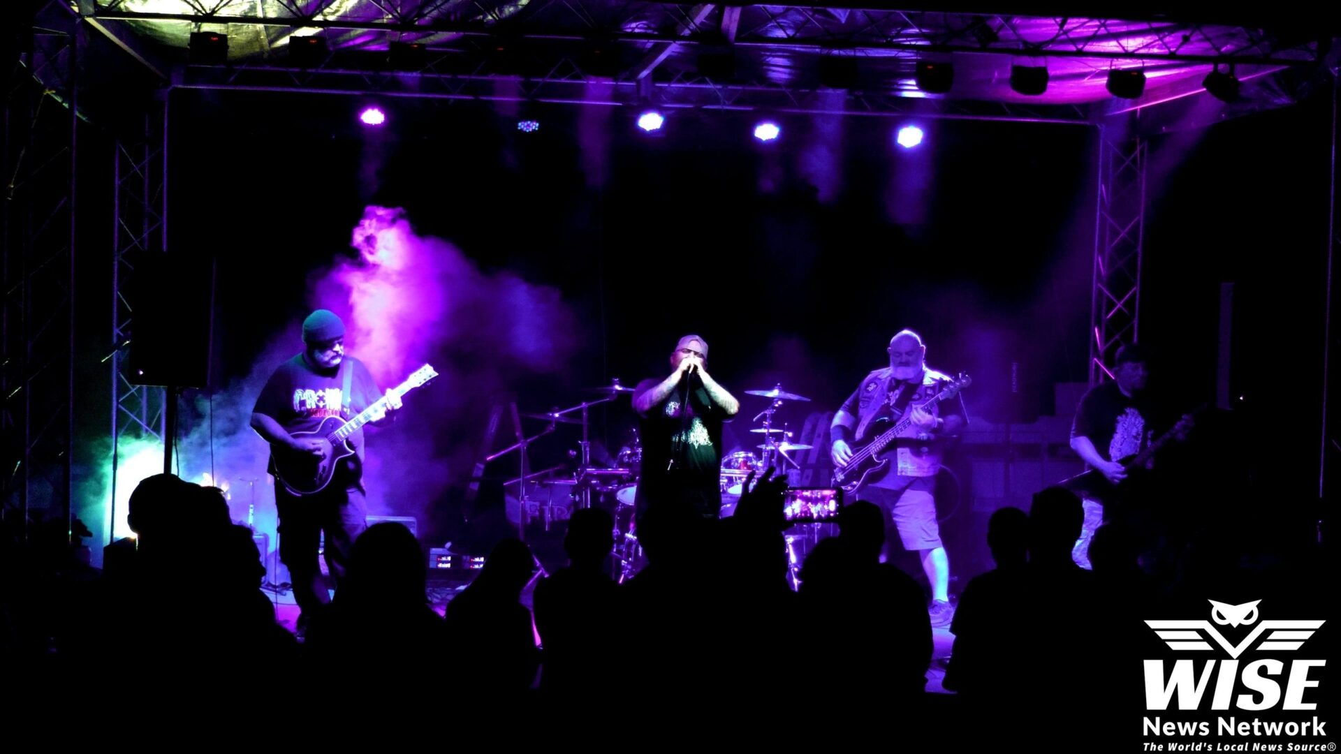 A band performs on a stage with vibrant purple and pink lighting, surrounded by instruments, as a crowd watches. The logo of "WISE News Network" is visible in the bottom right corner.