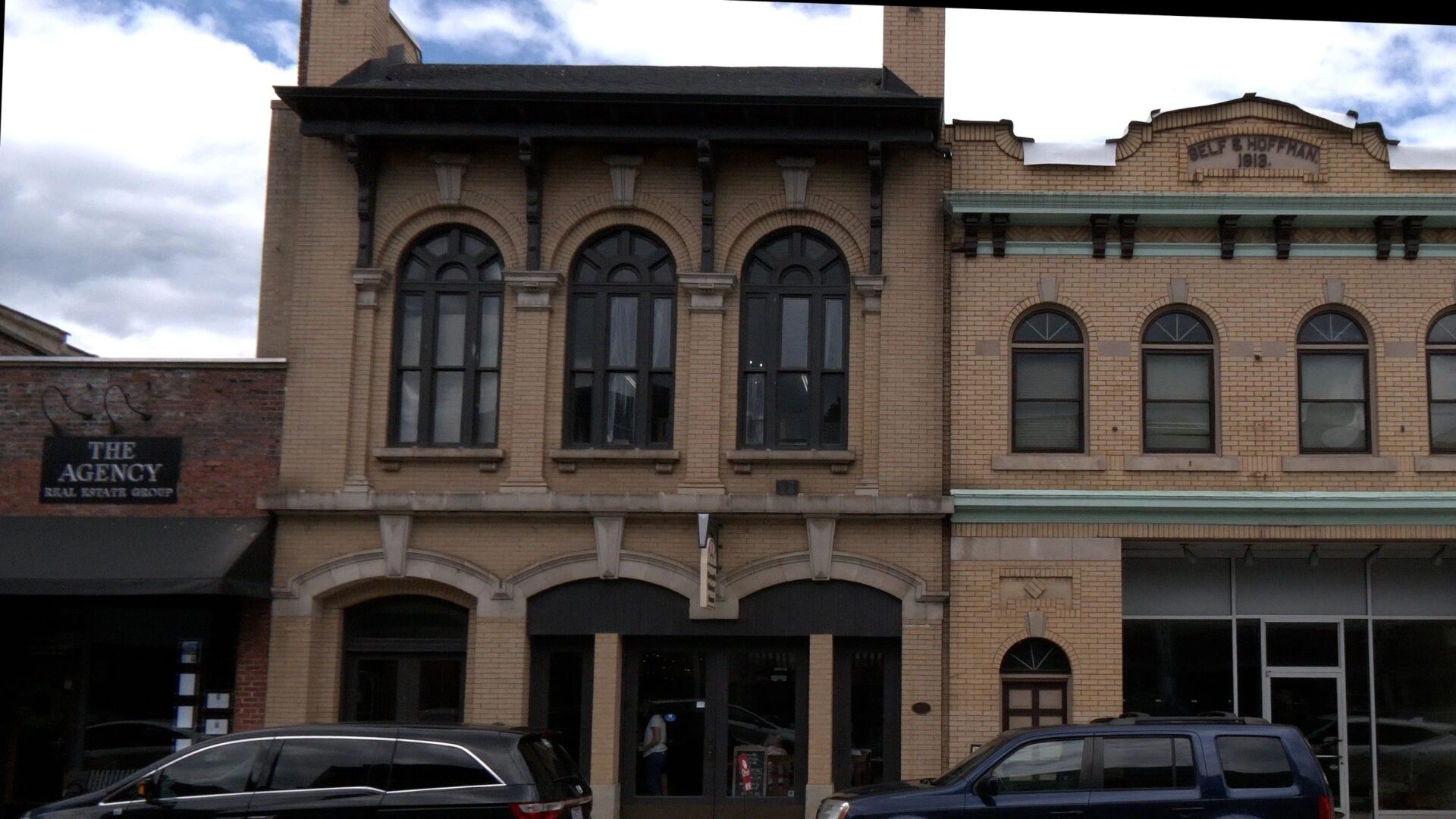 A historic two-story brick building with arched windows is flanked by adjacent structures, one of which displays a sign for a real estate agency. Vehicles are parked on the street in front.