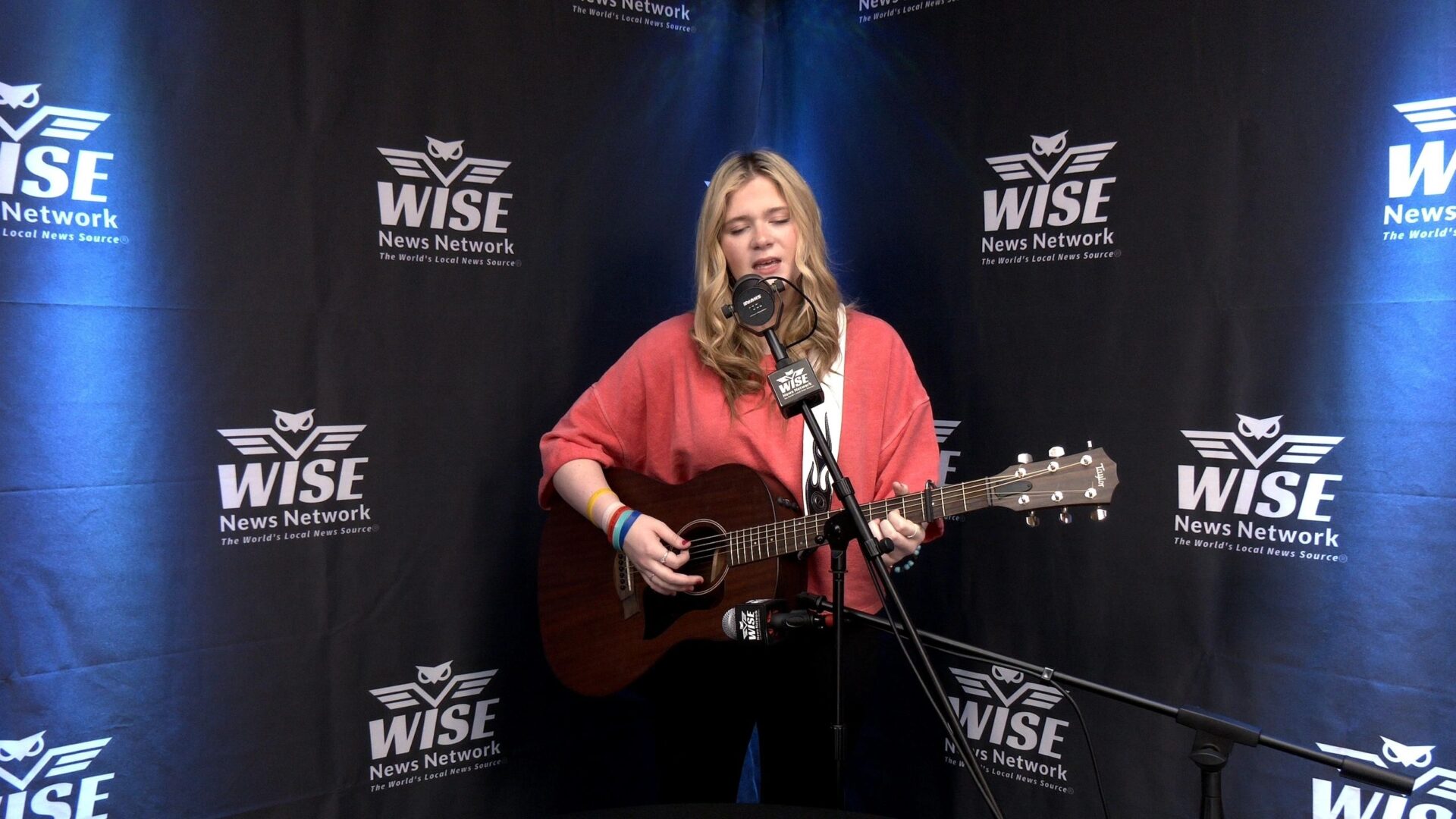 A person in a red sweater plays an acoustic guitar and sings into a microphone. They are standing in front of a backdrop with the WISE News Network logo.