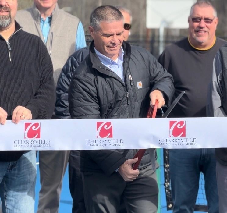 A group of people stands behind a ribbon being cut at a ceremony. The ribbon features the "Cherryville Chamber of Commerce" logo. One person in the center holds the large scissors.