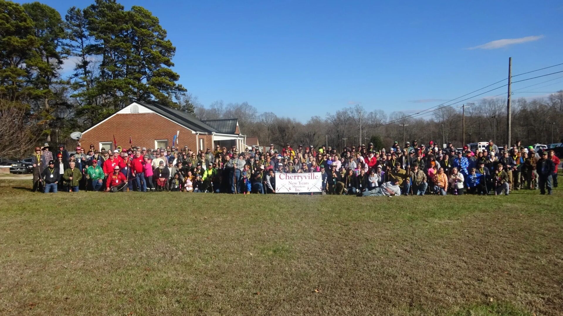 Large group of people standing together outside on a lawn in front of a brick building with a banner that reads "Cherryville" on a clear day.