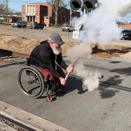 A person in a wheelchair is firing a small cannon on a railroad crossing. Smoke is rising from the cannon.