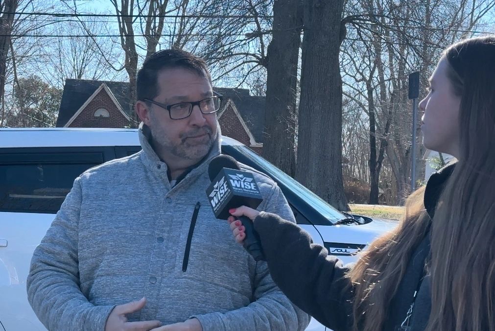 A man wearing glasses and a gray jacket is being interviewed by a woman holding a microphone labeled "WISX." They are standing outside near a white vehicle with trees and a house in the background.