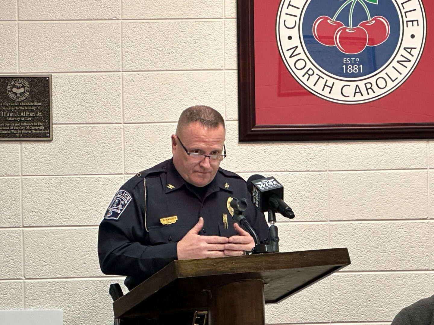 A police officer stands at a podium speaking into multiple microphones, with a wall plaque and a city emblem in the background.