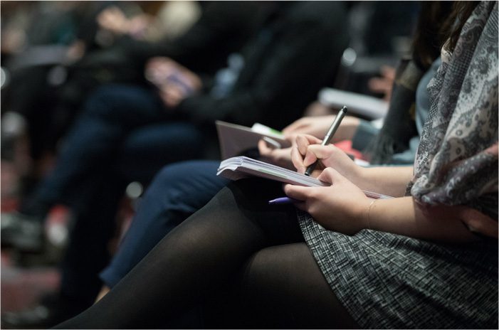 People seated and taking notes with pens and notebooks during what appears to be a conference or meeting.