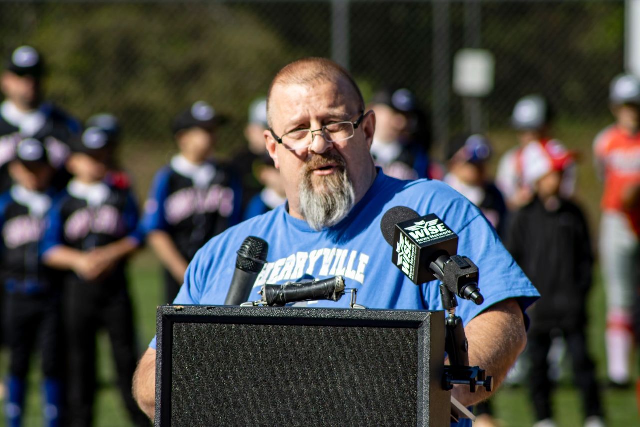 A man with glasses and a beard speaks at a podium with microphones. People in baseball uniforms stand in the background on a field.