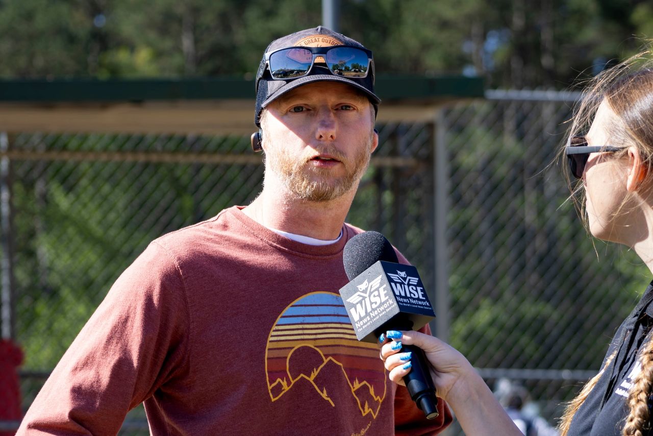 A man wearing sunglasses and a reddish-brown shirt is being interviewed by a woman holding a microphone. A chain-link fence and trees are visible in the background.