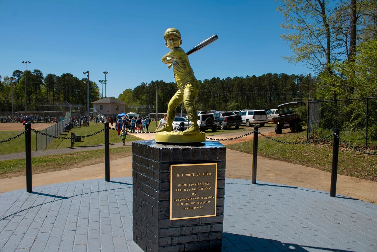 Golden statue of a baseball player in a batting stance on a brick pedestal with a plaque, located near a sports field and parking area, surrounded by a chain-link fence and forested background.