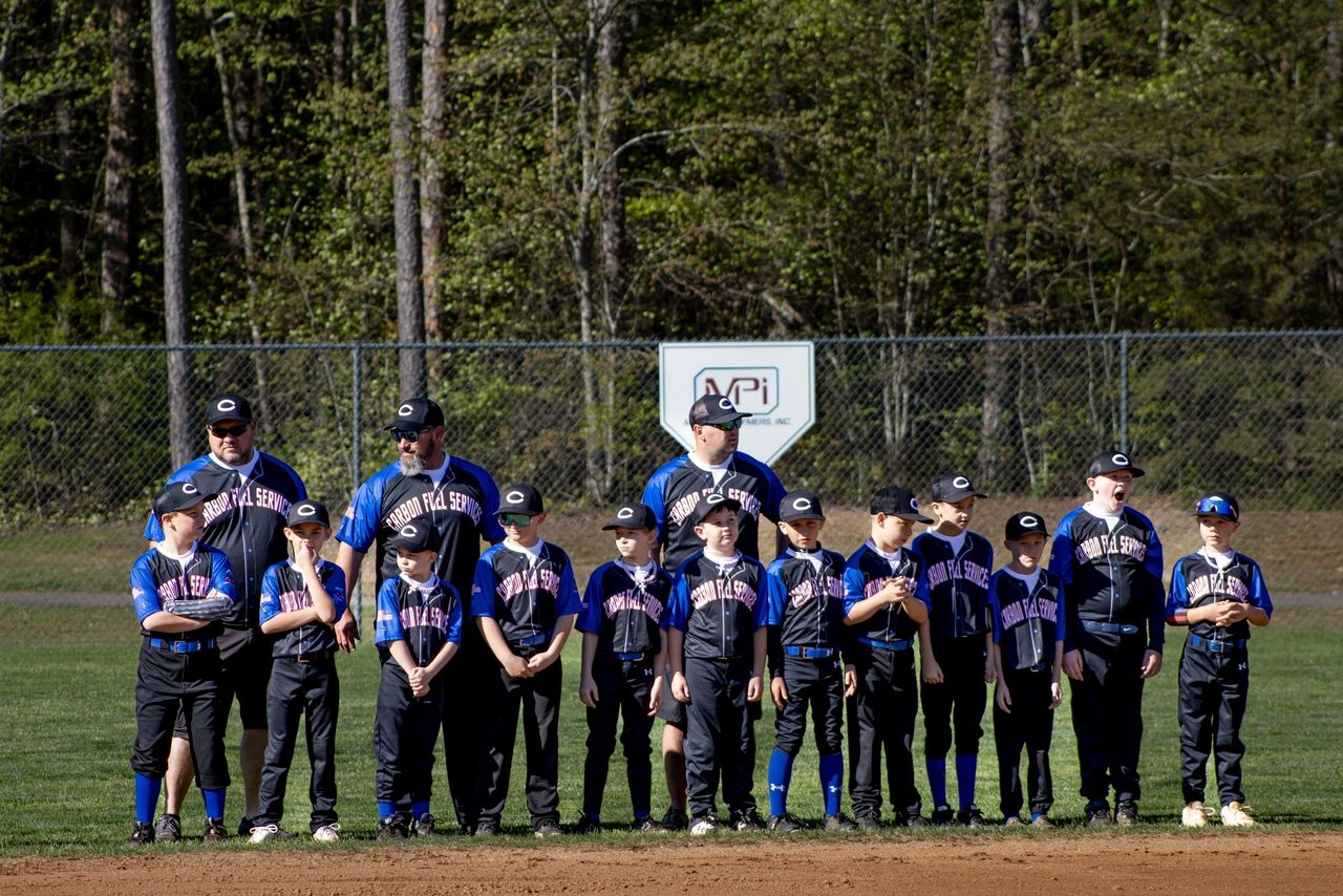 A baseball team lineup of young players and coaches, wearing blue and black uniforms, stands in front of a chain-link fence and trees during daytime.