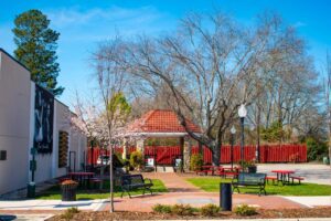 A small park with red picnic tables, green grass, and blossoming trees. A building with a red roof is in the background, and a mural is on the left wall. The sky is clear and blue.