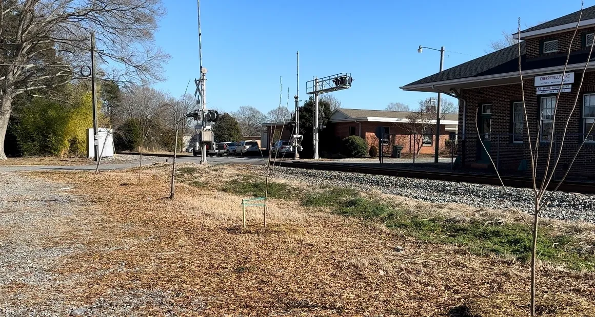 A railroad crossing with gates and lights. A brick building stands nearby. It's a clear day with trees in the background and sparse vegetation near the tracks.