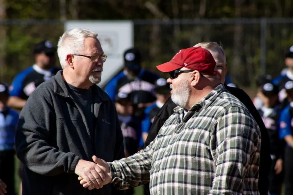 Two elderly men in casual clothing shake hands outdoors while others stand in the background.