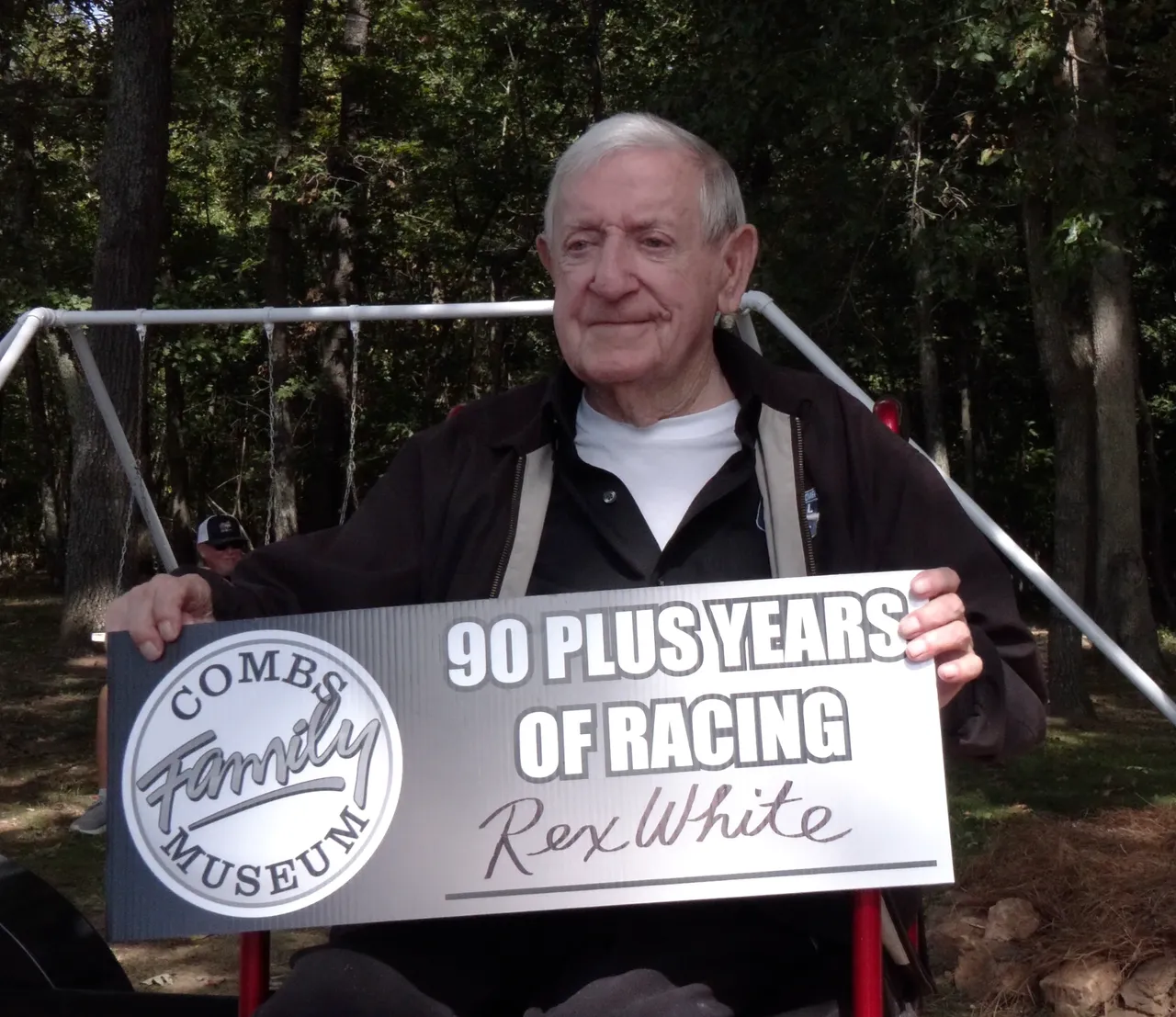 An elderly man is holding a sign that reads "90 Plus Years of Racing--Rex White" with a Combs Family Museum logo. He is seated outdoors with trees in the background.