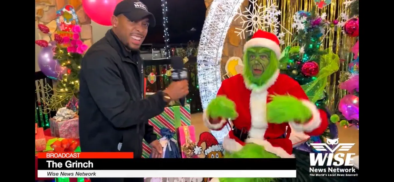 A man in a black jacket and cap interviews a person dressed as the Grinch in a festive white and red outfit, surrounded by Christmas decorations and gifts. Wise News Network branding is visible.