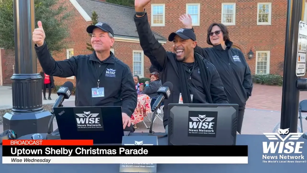 Three broadcasters wearing black Wise Network attire wave to the crowd during the Uptown Shelby Christmas Parade. They stand in front of laptops and microphones with a building in the background.