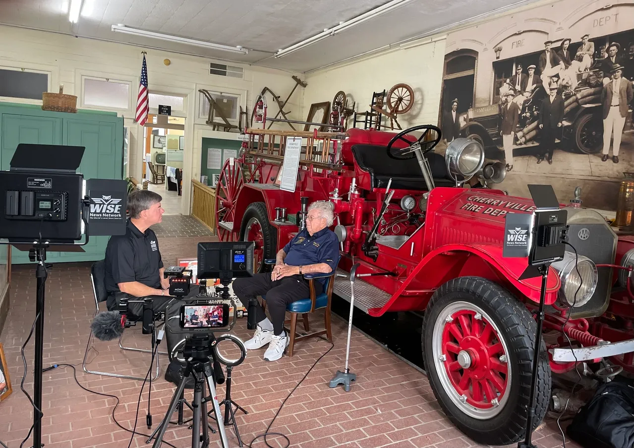 Two men are seated inside a museum with vintage fire trucks and equipment. There are cameras and lights set up, suggesting an interview or filming session. An American flag and historical photos are visible.