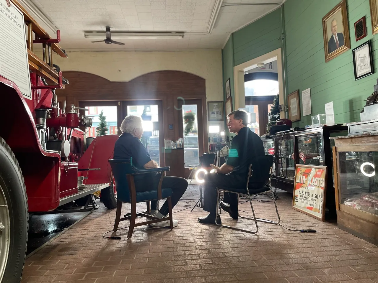 Two people are seated and engaged in conversation in a room featuring vintage items, including an old fire truck and framed photographs on the walls. Lighting and camera equipment are set up around them.