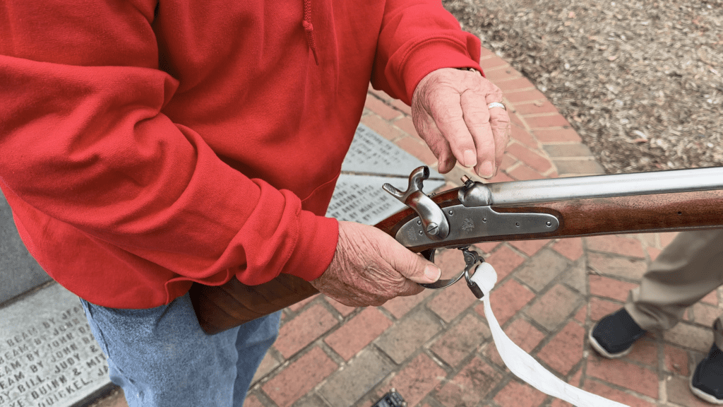 A man in a red hoodie holding a musket and putting a cap on to the firing pin of it. 