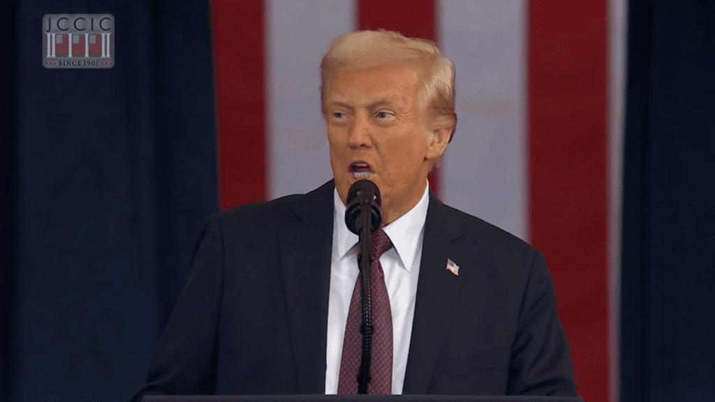 An older white male in a suit standing in front of an american flag.