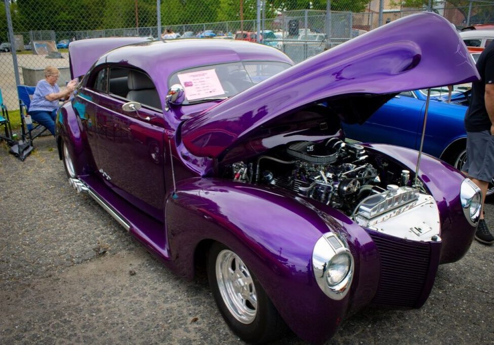 A purple classic car with an open hood displayed at a car show. An elderly person sits on a chair near the car, and several other vehicles are visible in the background.