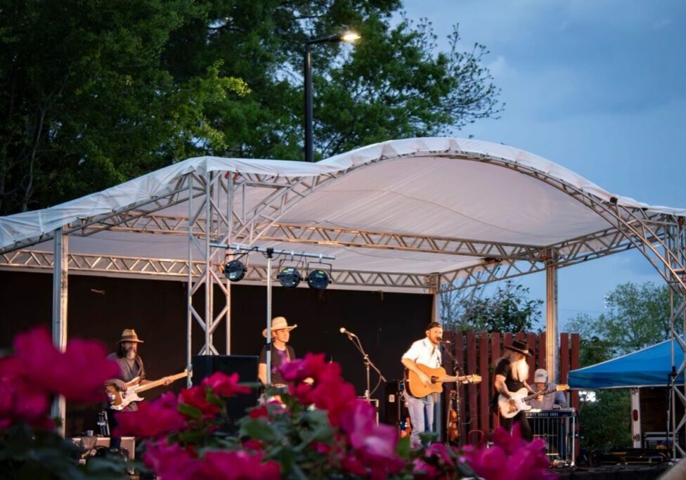 A band performs on an outdoor stage with a white canopy. Pink flowers are in the foreground, and trees and evening sky are in the background.