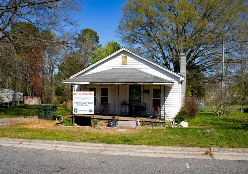 Small white house with a "No Trespassing" sign and multiple trash bins outside. Surrounded by trees, it has a porch with various items on it.