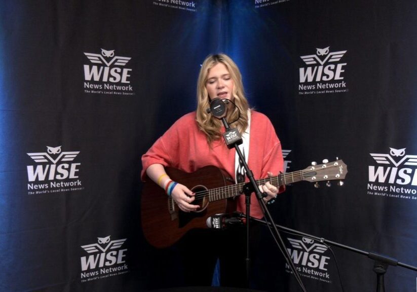 A woman with long blonde hair is playing an acoustic guitar and singing into a microphone in a studio with WISE News Network banners in the background.