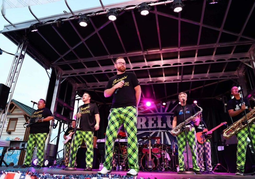 A band performs on an outdoor stage, wearing matching green plaid pants and black t-shirts. The stage is lit with purple lights and various musical instruments are visible.