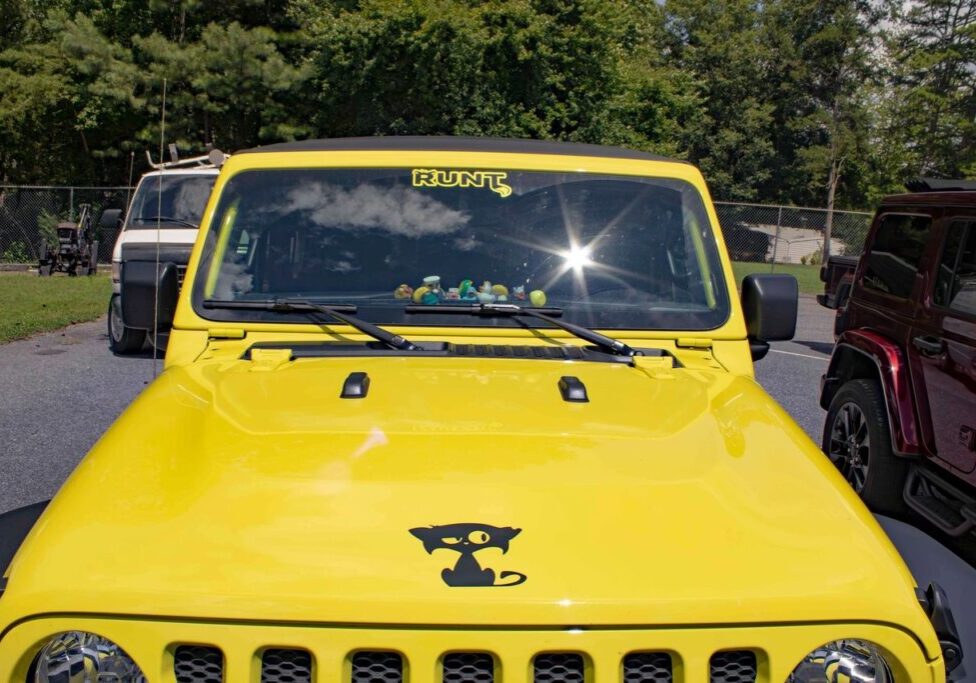 A bright yellow Jeep with a black cat decal on the hood and character figurines on the dashboard is parked in a lot with trees and other vehicles in the background.