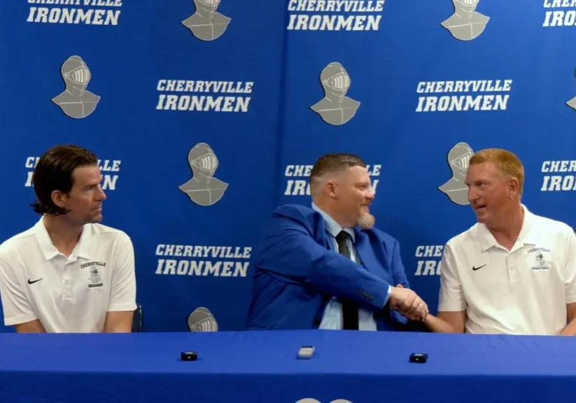 Three men are seated at a table in front of a backdrop labeled "Cherryville Ironmen." The man in the middle, wearing a blue suit, shakes hands with the man on the right, while the man on the left observes.