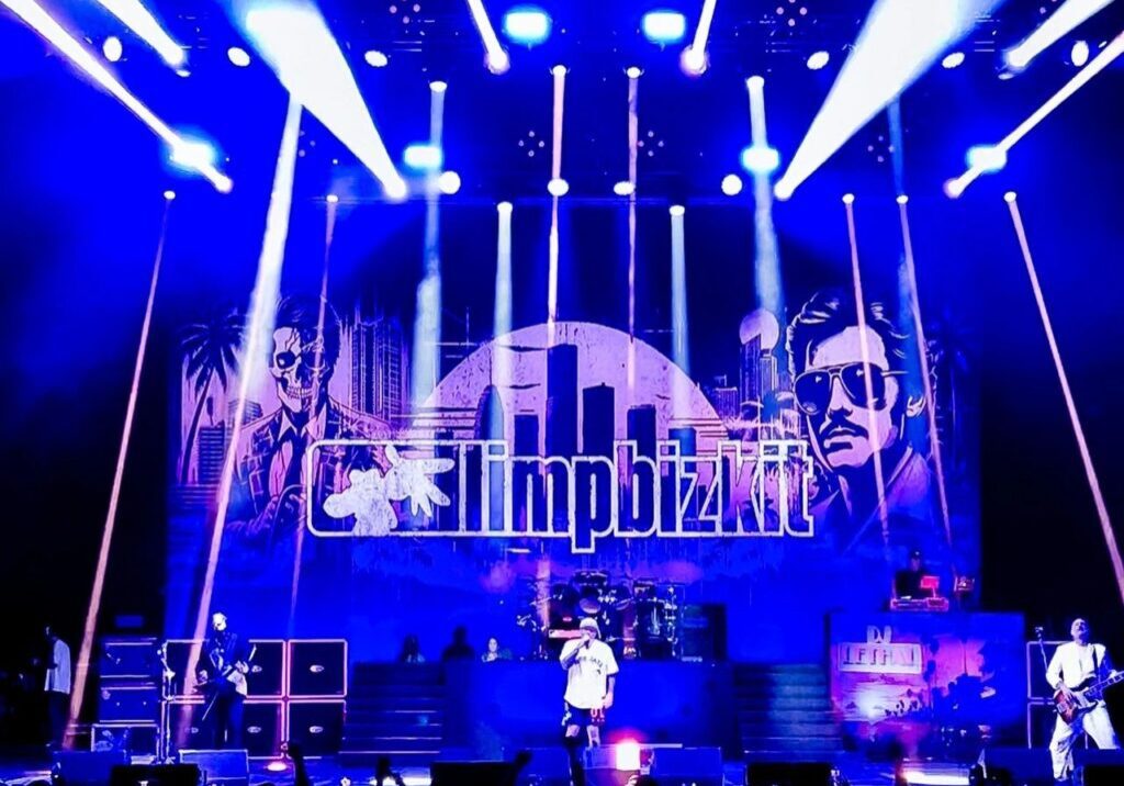 A band performs on a stage with bright lights and a backdrop displaying "limpbizkit" in blue and purple hues, featuring two illustrated faces.