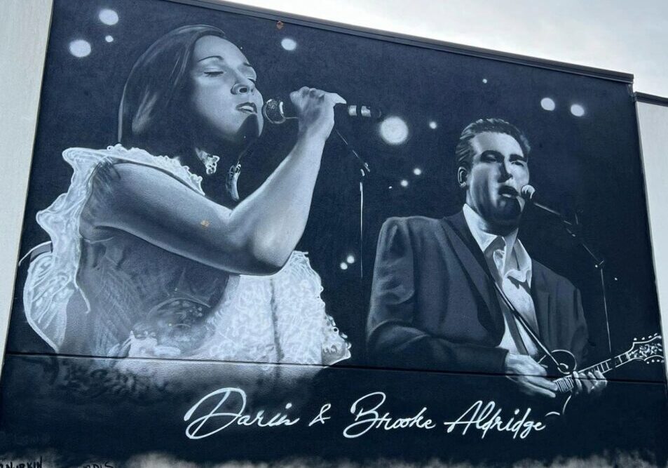 A mural painted on a wall depicts a female and a male singer performing on stage. The mural includes their signed names.