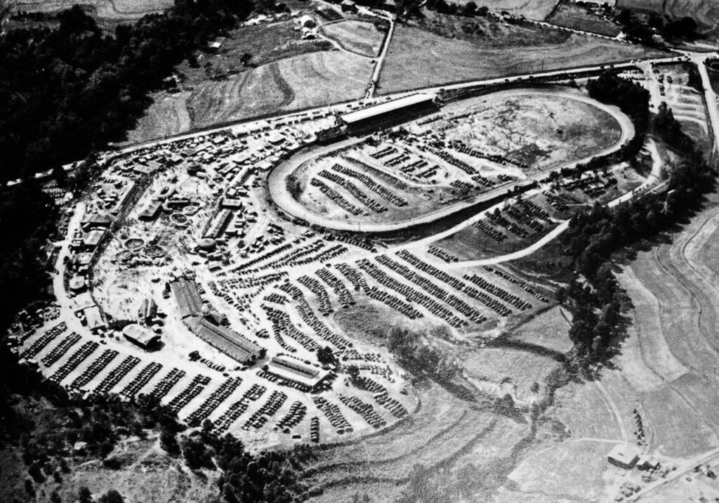 Aerial view of a large outdoor event displaying an oval race track, numerous parked vehicles, various buildings, and surrounding fields and trees.