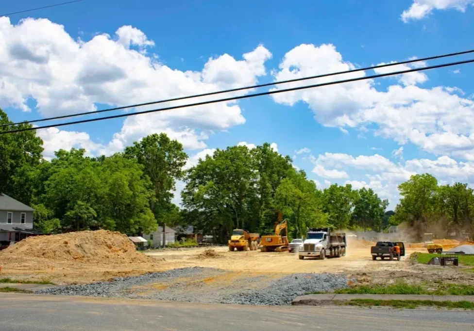 Construction site with heavy machinery including excavators and dump trucks on a lot cleared of vegetation. Piles of dirt and gravel are visible around the site. Bright, sunny day with blue sky and clouds.