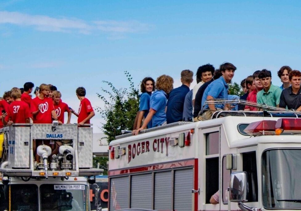 A group of people, mostly young men, stand on top of two fire trucks labeled "Boger City" and "Dallas." Some are wearing red and blue shirts. Trees and a blue sky are in the background.