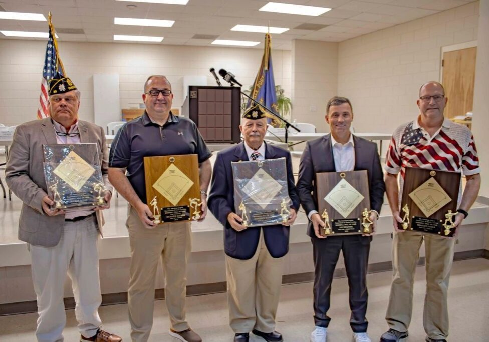 Five men standing indoors, each holding a plaque with trophies, in front of a podium and flags.